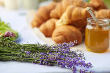 Close up of table decorated with fresh delicious croissants, honey jar with wooden spoon and aromatic purple lavender bouquet. Beautiful serving outdoors. Concept of cuisine, pastry. © serhiibobyk