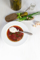 Borshct with sour cream in bowl, vodka glass, garlic, onion, salo, bread, glass jar with pickled cucumbers on white background