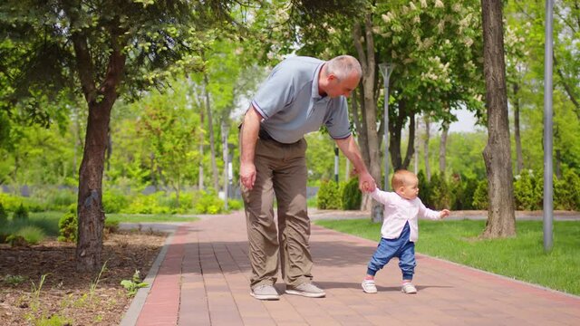 Baby learns to walk outdoors. Grandpa supports his granddaughter teaching to walk. baby girl takes first steps.