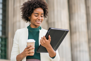 Afro businesswoman holding a cup of coffee and clipboard outdoors.