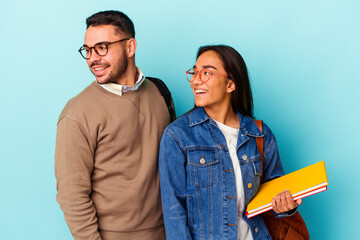 Young mixed race student couple isolated on blue background looks aside smiling, cheerful and pleasant.