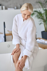 Happy smiling gorgeous senior lady in bath robe at spa center sitting on bathtube touching legs. Advertising of bodycare spa procedures antiage recreation skin care salon concept. Vertical shot.