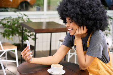 Fototapeta na wymiar Young beautiful mixed race cheerful student hipster girl talking on mobile app on video conference sitting in outdoor cafe using cellphone. Blogger influencer recording video for followers.