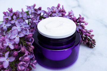 Close-up of a purple jar with natural cosmetic cream and a branch of lilac on a light background.