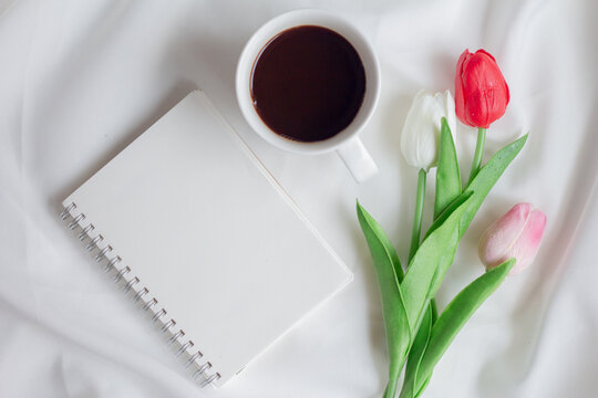 Blank paper notebook with cup of coffee and tulip flowers on white fabric. Top view, flat lay.