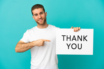 Handsome blonde man over isolated blue background holding a placard with text THANK YOU and  pointing it