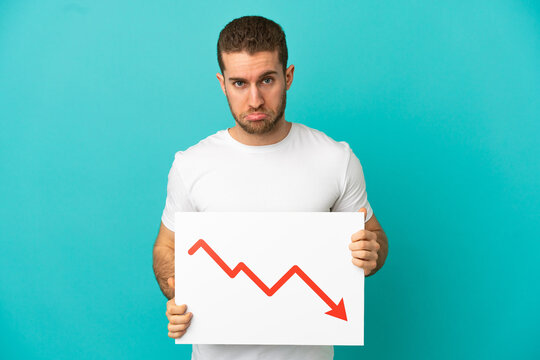 Handsome blonde man over isolated blue background holding a sign with a decreasing statistics arrow symbol with sad expression