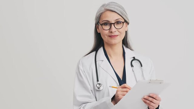 A pleased senior woman doctor is holding a folder with documents standing isolated over grey wall in the studio