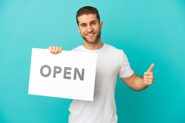 Handsome blonde man over isolated blue background holding a placard with text OPEN with thumb up