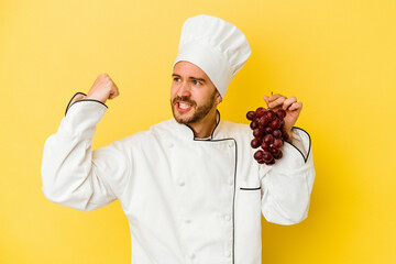 Young caucasian chef man holding grapes isolated on yellow background raising fist after a victory, winner concept.