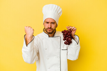 Young caucasian chef man holding grapes isolated on yellow background showing fist to camera, aggressive facial expression.