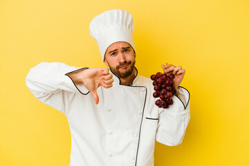 Young caucasian chef man holding grapes isolated on yellow background showing a dislike gesture, thumbs down. Disagreement concept.