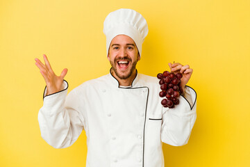 Young caucasian chef man holding grapes isolated on yellow background receiving a pleasant surprise, excited and raising hands.