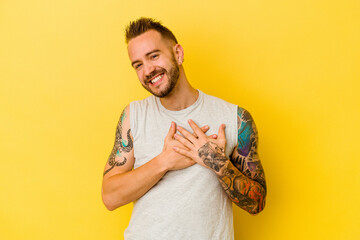 Young tattooed caucasian man isolated on yellow background laughing keeping hands on heart, concept...
