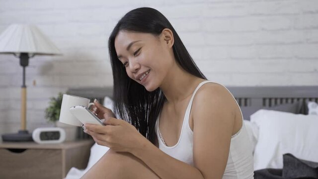portrait gorgeous asian female holding coffee and a cell phone is chuckling while enjoying funny posts on social media at home.
