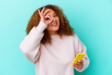 Young latin woman holding mobile phone isolated on blue background excited keeping ok gesture on eye.