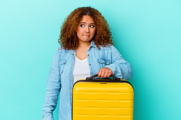 Young latin traveler curvy woman holding a suitcase isolated on blue background confused, feels doubtful and unsure.