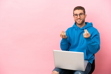 Young man sitting on a chair with laptop making money gesture
