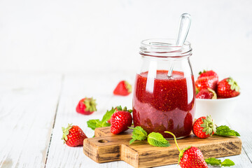 Strawberry jam in the glass jar at white table with fresh strawberries.