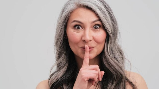 A surprised elder woman is showing secret gesture standing isolated over grey wall in the studio