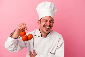 Young caucasian chef man holding tomatoes isolated on pink background laughing and having fun.