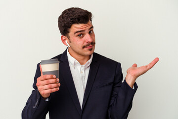 Young business caucasian man wearing wireless headphones and holding take way coffee isolated on white background showing a copy space on a palm and holding another hand on waist.