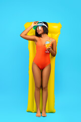 Full length of relaxed black lady in swimsuit lying on yellow inflatable lilo and drinking cocktail over blue background - obrazy, fototapety, plakaty