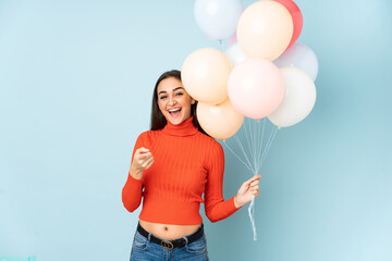 Young woman catching many balloons isolated on blue background points finger at you