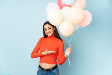 Young woman catching many balloons isolated on blue background extending hands to the side for inviting to come