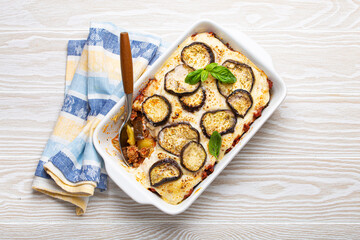 Greek mediterranean dish Moussaka with baked eggplants, ground beef in white ceramic casserole with napkin on wooden white background from above, traditional dish of Greece