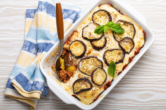 Greek Mediterranean Dish Moussaka With Baked Eggplants, Ground Beef In White Ceramic Casserole With Napkin On Wooden White Background From Above, Traditional Dish Of Greece