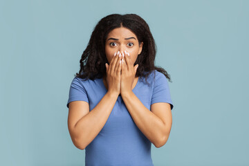 Oh no. Shocked and scared african american lady covering her mouth with palms, standing over blue...