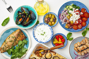 Assorted Greek dishes on white stone background from above, moussaka, grilled fish, souvlaki, greek salad, steamed mussels with herbs, appetizers of Greece from above 