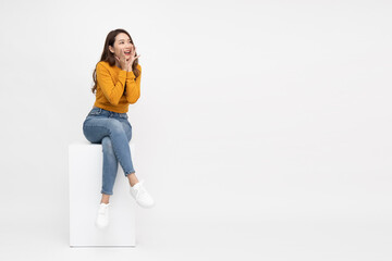 Portrait of excited screaming young asian woman sitting on white box isolated over white...