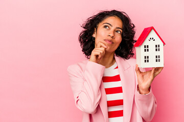 Young latin woman holding a toy house isolated on pink background looking sideways with doubtful...