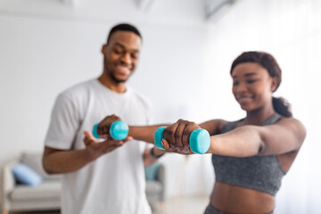 Personal trainer showing young black lady how to exercise with dumbbells correctly at home, selective focus