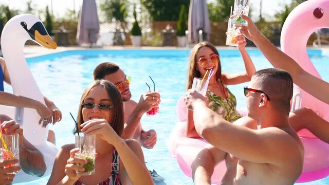 Friends having party toasting with cocktails in holiday villa swimming pool. Happy young people in swimwear dancing, clubbing with inflatable flamingo, swan, at resort on sunny day. Clinking glasses.