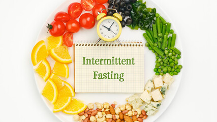 Circle of assorted fresh food on a plate. Clock and notebook with the text Intermittent Fasting....