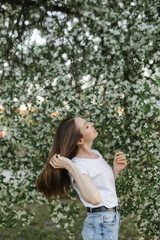 Caucasian girl with brown hair in blue jeans and a white T-shirt on the background of an apple tree in bloom. Sunset. The concept of spring flowering. Apple tree in bloom.