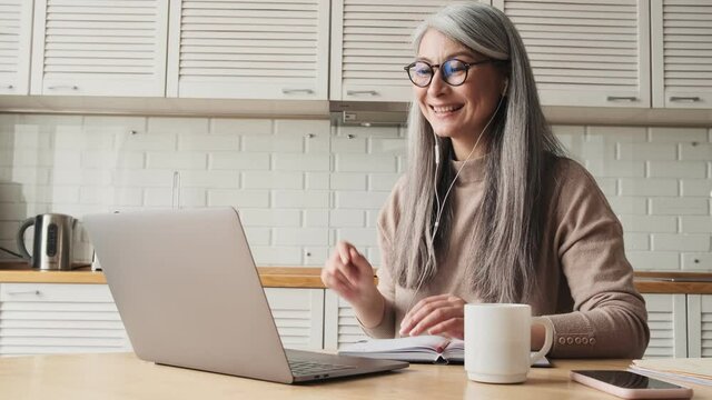 A smiling elder woman is talking with video-connection using her laptop sitting in the kitchen at home