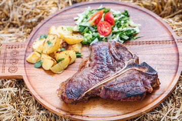 T bone steak served on a wooden board placed on a straw, with salad, potatoes 