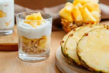 Delicious pineapple dessert. Breakfast dessert with oat granola, greek yogurt and pineapple in layers in glass with ingredients chopped fresh juicy pineapple on wooden table.