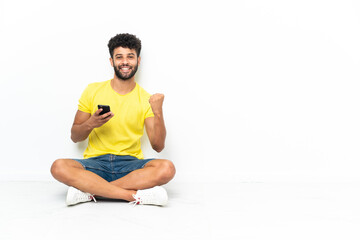 Fototapeta premium Young Moroccan handsome man sitting on the floor over isolated background with phone in victory position