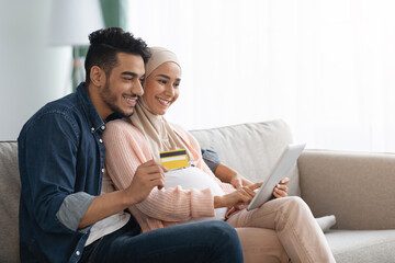 Cheerful pregnant muslim couple shopping online, using credit card and digital tablet