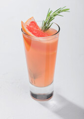 Glass of fresh pressed ruby red grapefruit juice with alcohol on white background with rosemary and fruit slice.