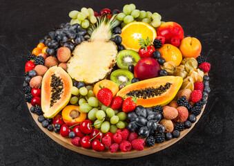 Naklejka na ściany i meble Fresh raw organic summer berries and exotic fruits in round large tray on black kitchen background. Papaya, grapes, nectarine, orange, raspberry, kiwi, strawberry, lychees, cherry and pineapple.
