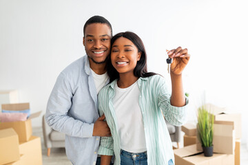 Real estate. Happy african american couple showing new house key, standing against cardboard boxes
