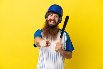 Redhead baseball player man with helmet and bat isolated on yellow background surprised and pointing front