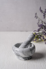 On a white textured background is a granite mortar for grinding spices. The kitchen mortar is made of white marble. In the background, Provencal herbs. Natural materials. Cooking at home.