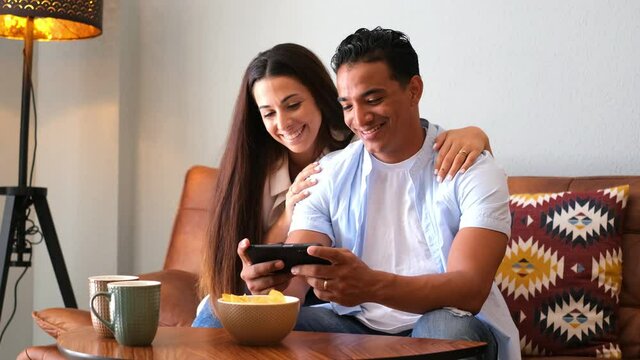 Young happy couple use smart phone together at home sitting on the couch - cheerful girl and boy interracial people using celllular to message and have fun - love and life relationship concept